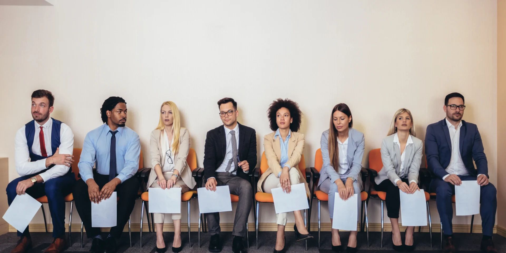 A diverse group of job applicants sitting in a row, holding resumes and waiting for interviews.