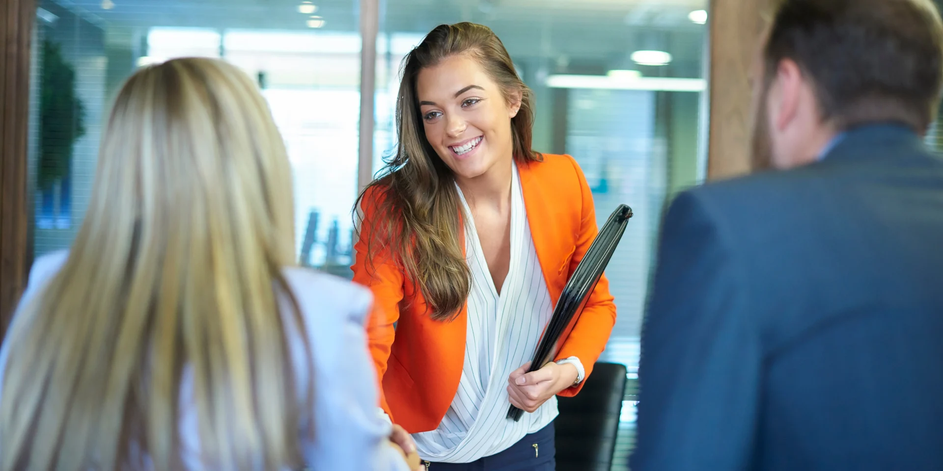 A confident woman in a bright orange blazer shaking hands with an interviewer during a job interview.