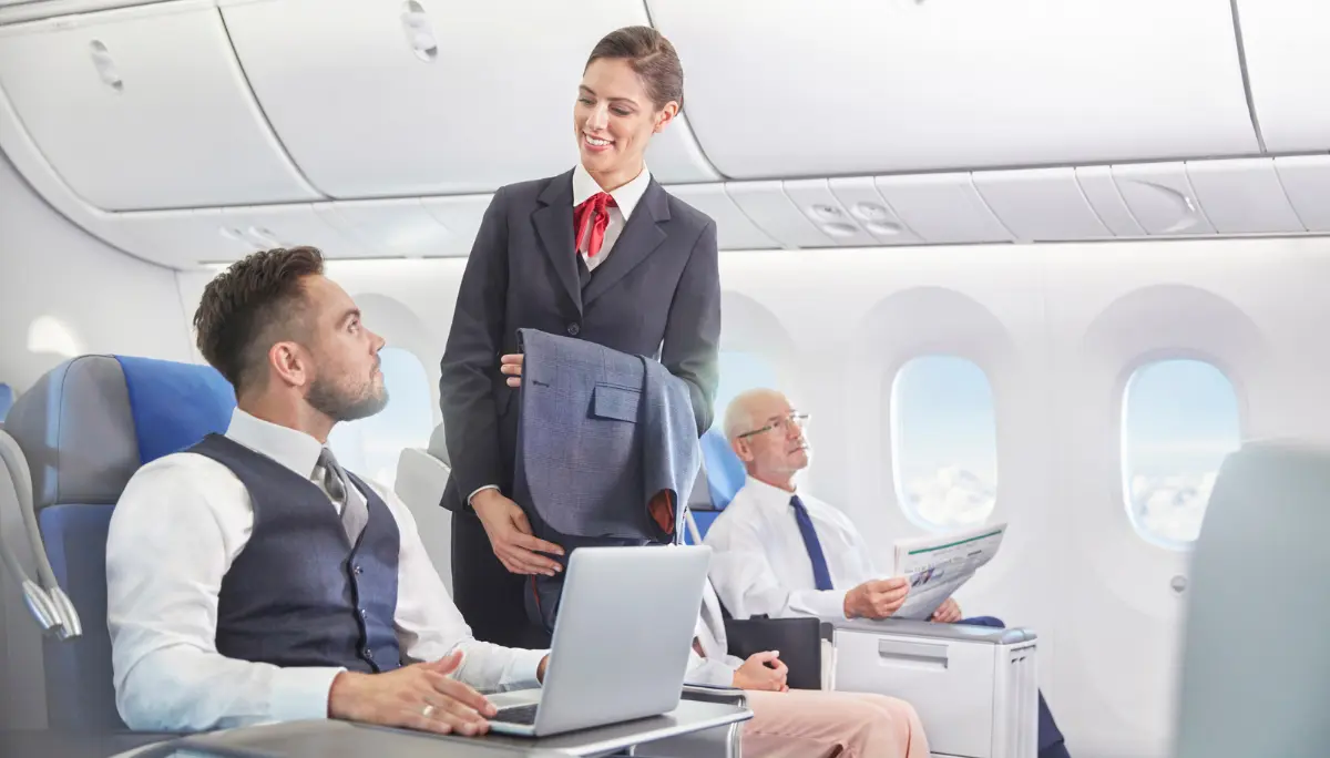 Professionalism in Premium Cabins Flight attendant in a blazer hands a jacket to a male passenger in business class, while another passenger reads nearby.