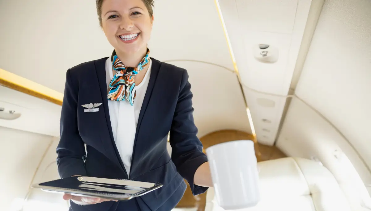 Business-Class Service with First-Class Composure Professional flight attendant in a private jet cabin smiling and holding a coffee mug and tray.