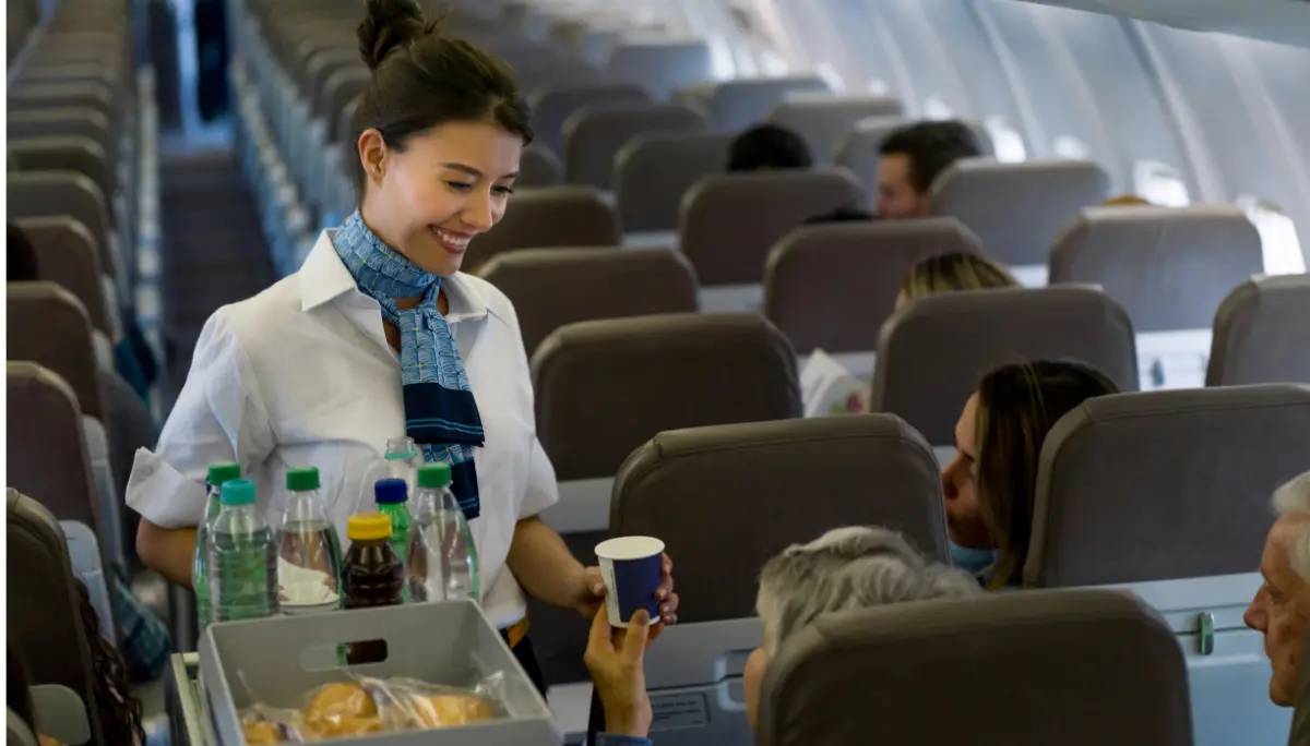 Smiling flight attendant hands a cup to a passenger while pushing a beverage cart down the aisle.