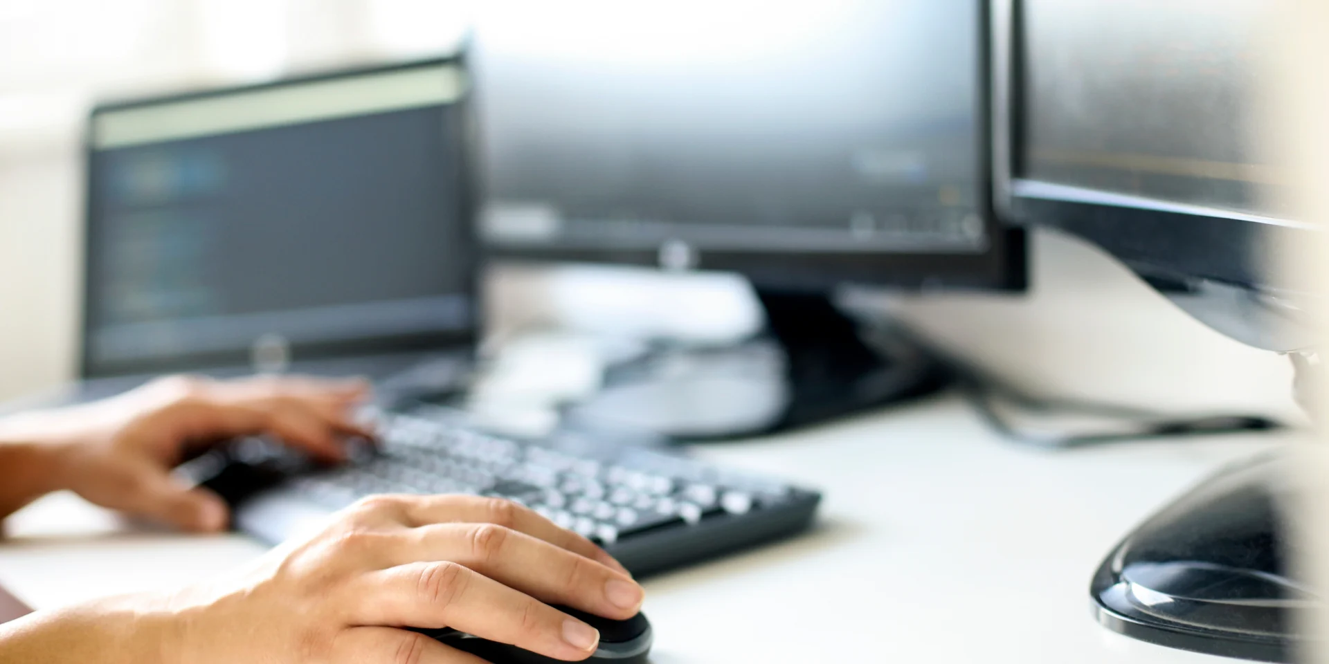 A close-up view of a person's hands on a keyboard and mouse, working at a desk with multiple computer monitors, representing active and focused interview practice.